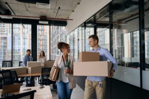 Deux coworkers souriants portent des boîtes et du matériel de bureau dans un espace de coworking moderne, prêt à déménager, illustrant bien préparer son déménagement fiche pratique.