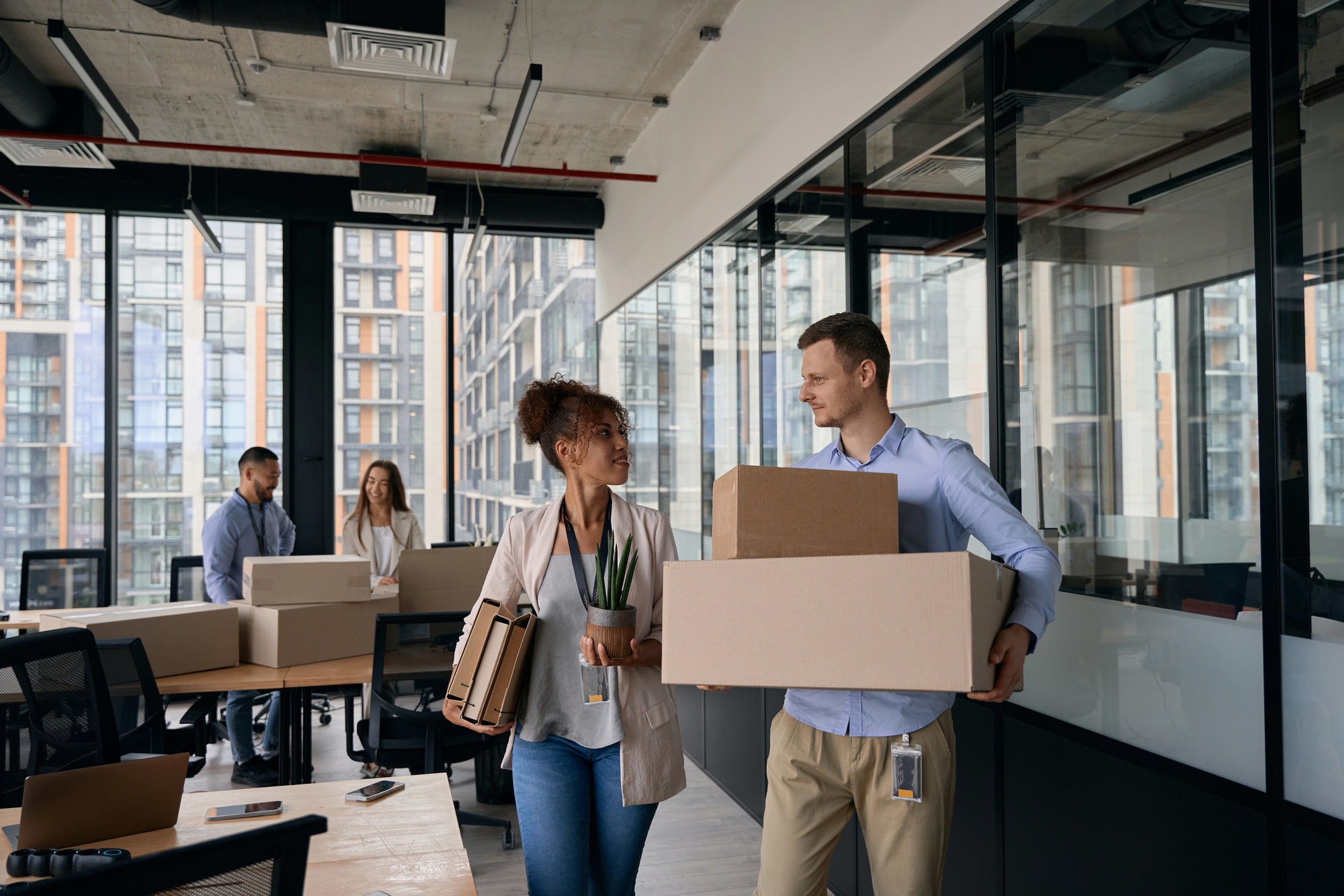 Deux coworkers souriants portent des boîtes et du matériel de bureau dans un espace de coworking moderne, prêt à déménager, illustrant bien préparer son déménagement fiche pratique.
