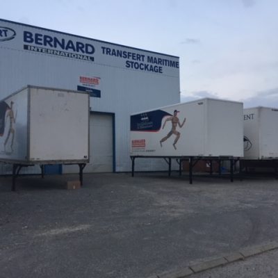 Trains routiers et remorques stationnées devant un entrepôt portant le texte Bernard Transfert Maritime Stockage.